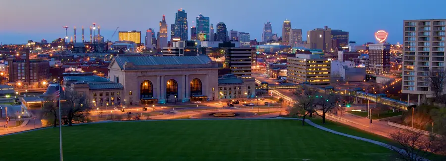 Downtown Kansas City at dusk: the lit skyline rises behind Union Station, with traffic trails on the road and the Liberty Memorial lawn in the foreground.