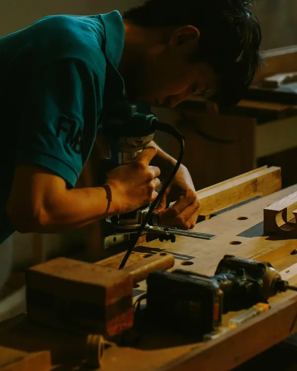 A carpenter in a teal shirt guides a router along a jig on a wooden workbench in warm workshop light.